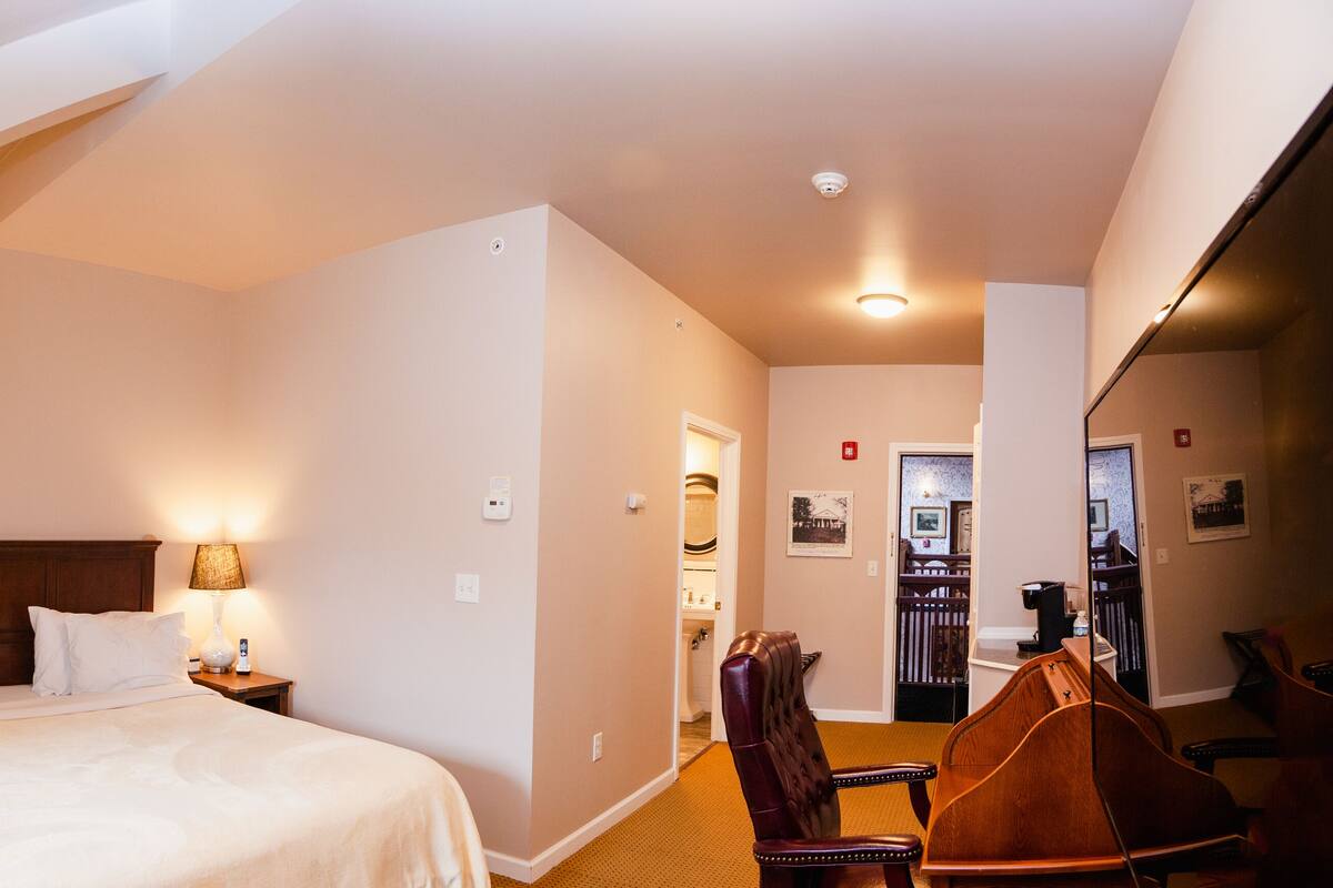 Ein Hotelzimmer mit einem großen Bett, zwei Nachttischlampen, einem Holz-Kopfteil, einem Holzsessel und einem Schreibtisch mit Stuhl.