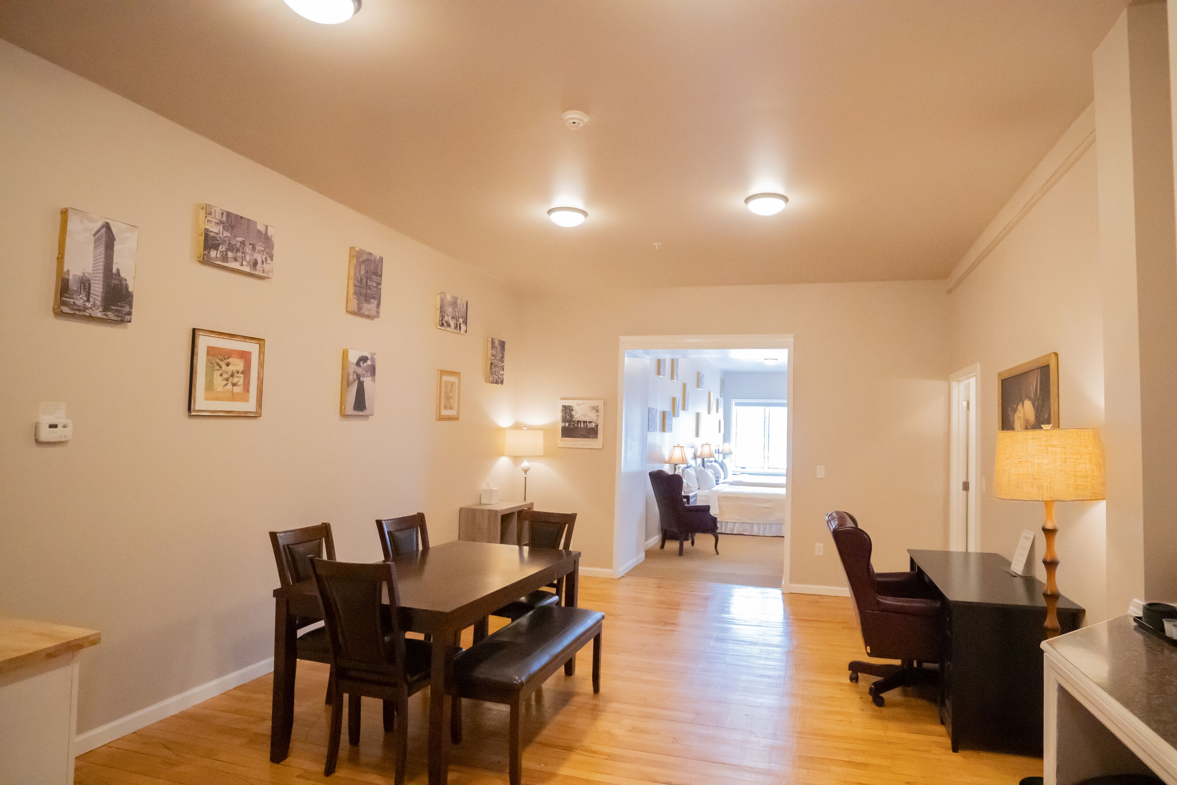 A dining area with a wooden table and chairs, a bench, and a desk with a lamp. There are framed pictures on the walls and a hallway leading to another room.