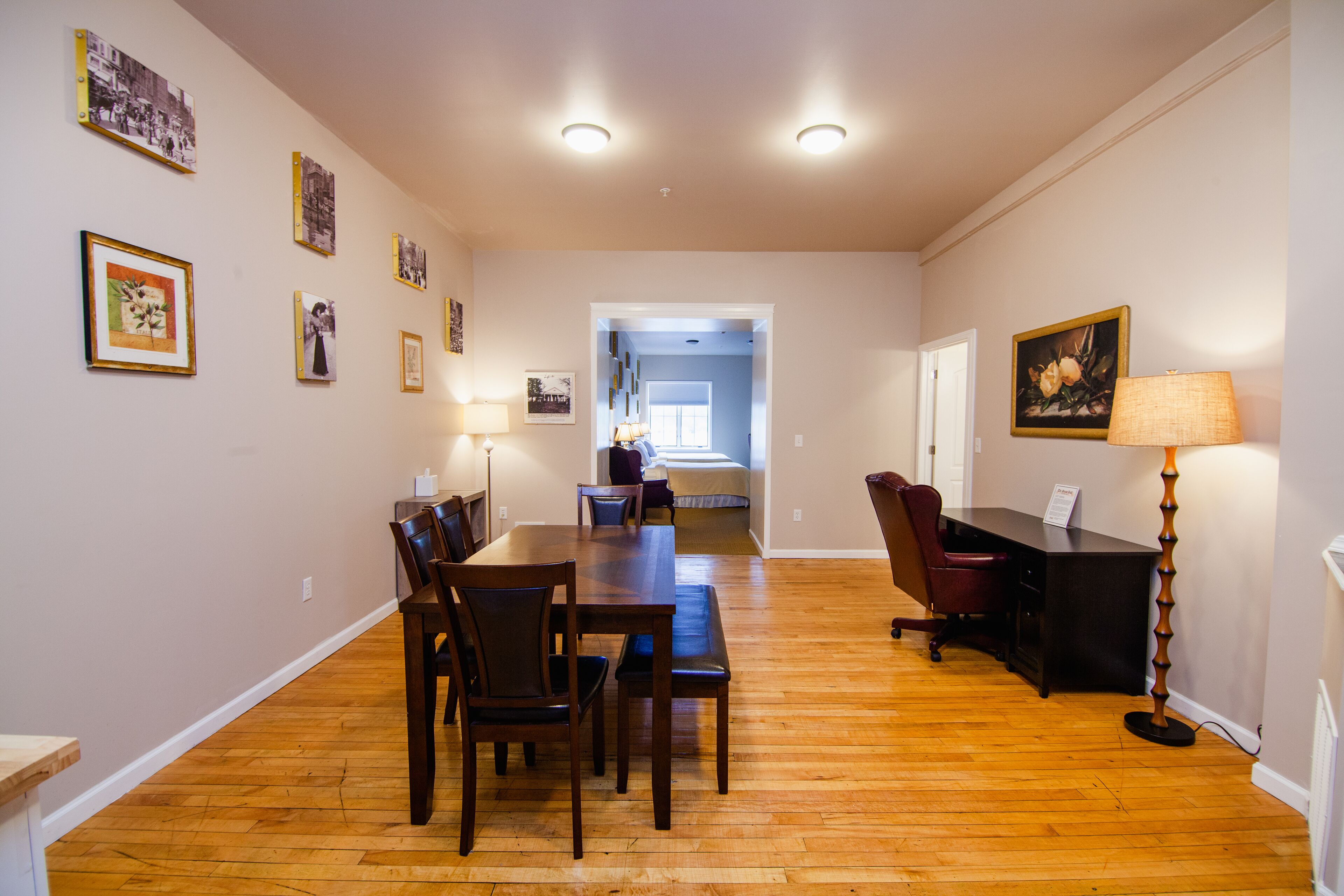 Una habitación amplia con una mesa de comedor de madera, sillas, un escritorio y una lámpara. Se observan cuadros en las paredes.