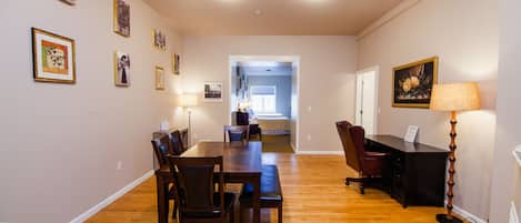 A spacious room with a wooden dining table, chairs, a desk, and a lamp. There are framed pictures on the walls.