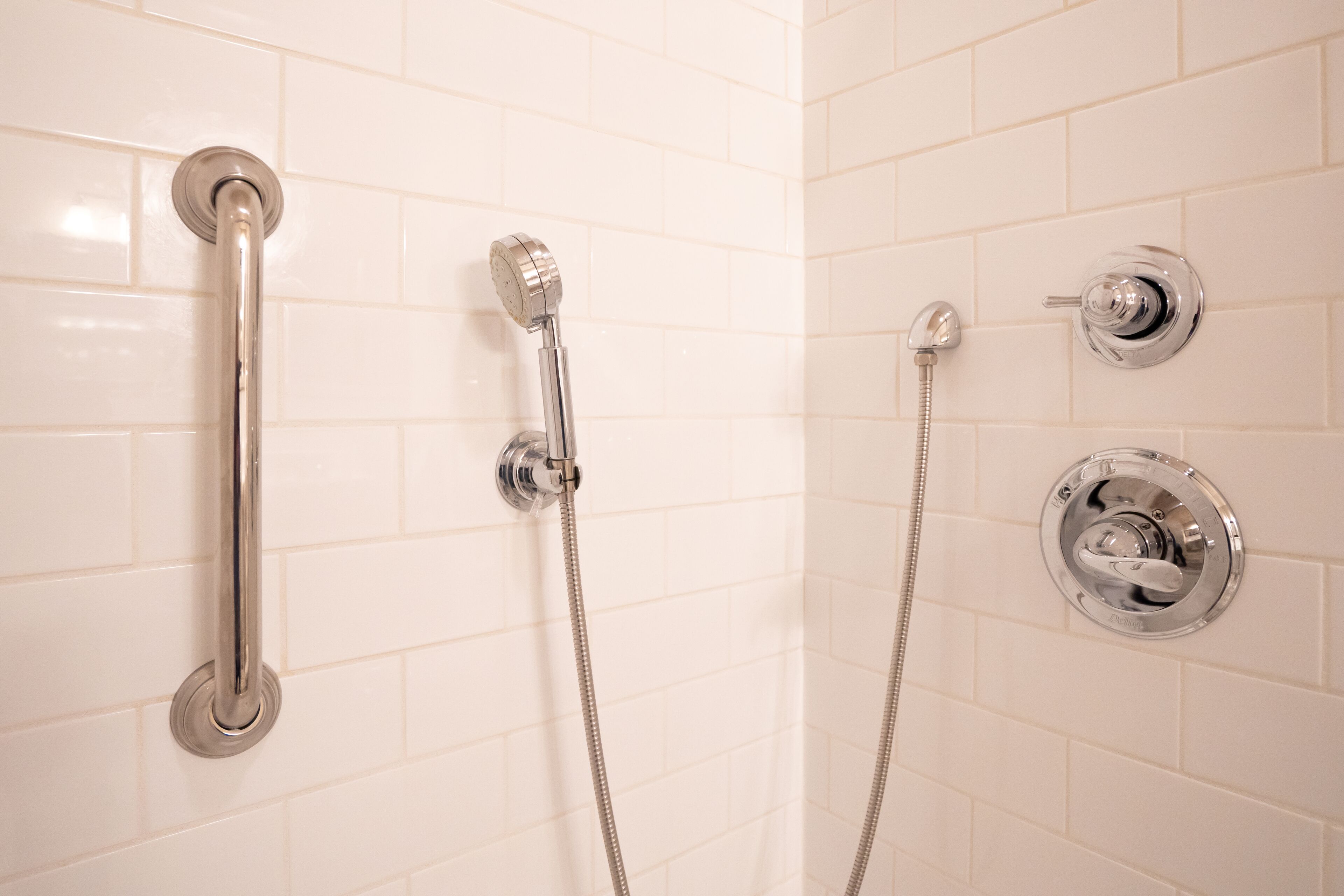 A bathroom with white tiled walls, a chrome showerhead, a handheld showerhead, and a wall-mounted faucet.