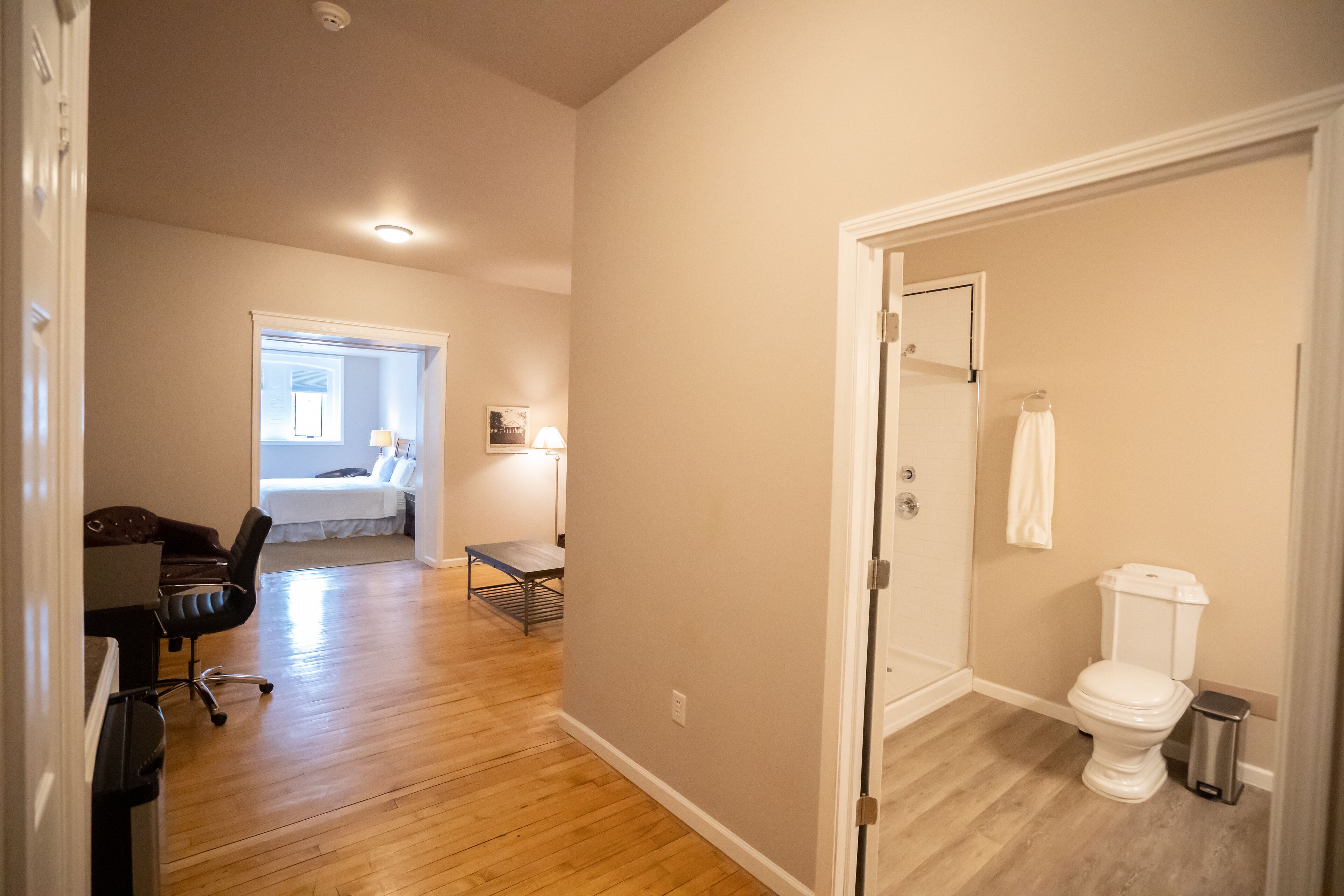 A room with a wooden floor, a desk with a chair, a bed, a small table with a lamp, and a toilet.