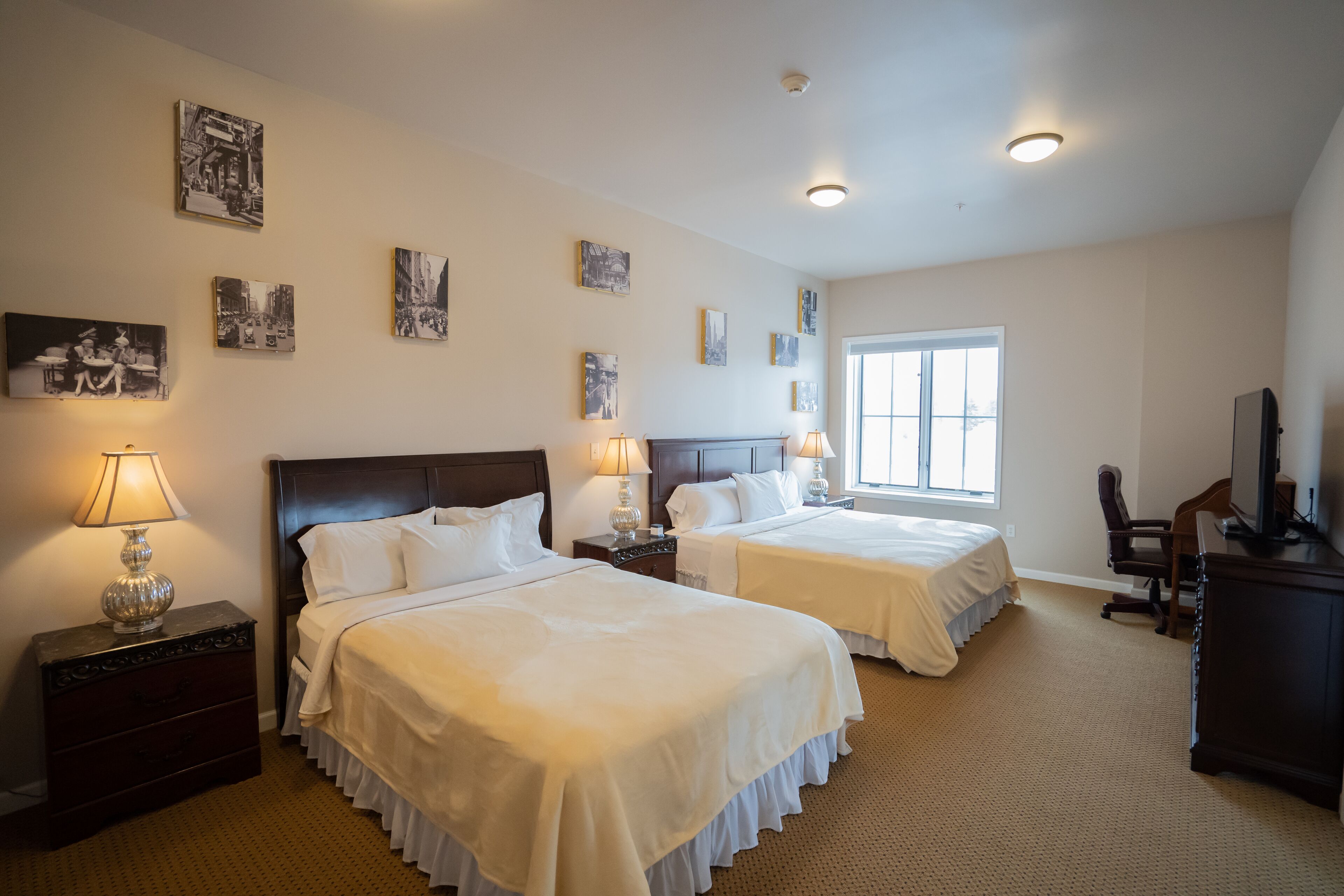 Quarto de hotel com duas camas, uma escrivaninha, uma cadeira, televisão e obras de arte nas paredes.