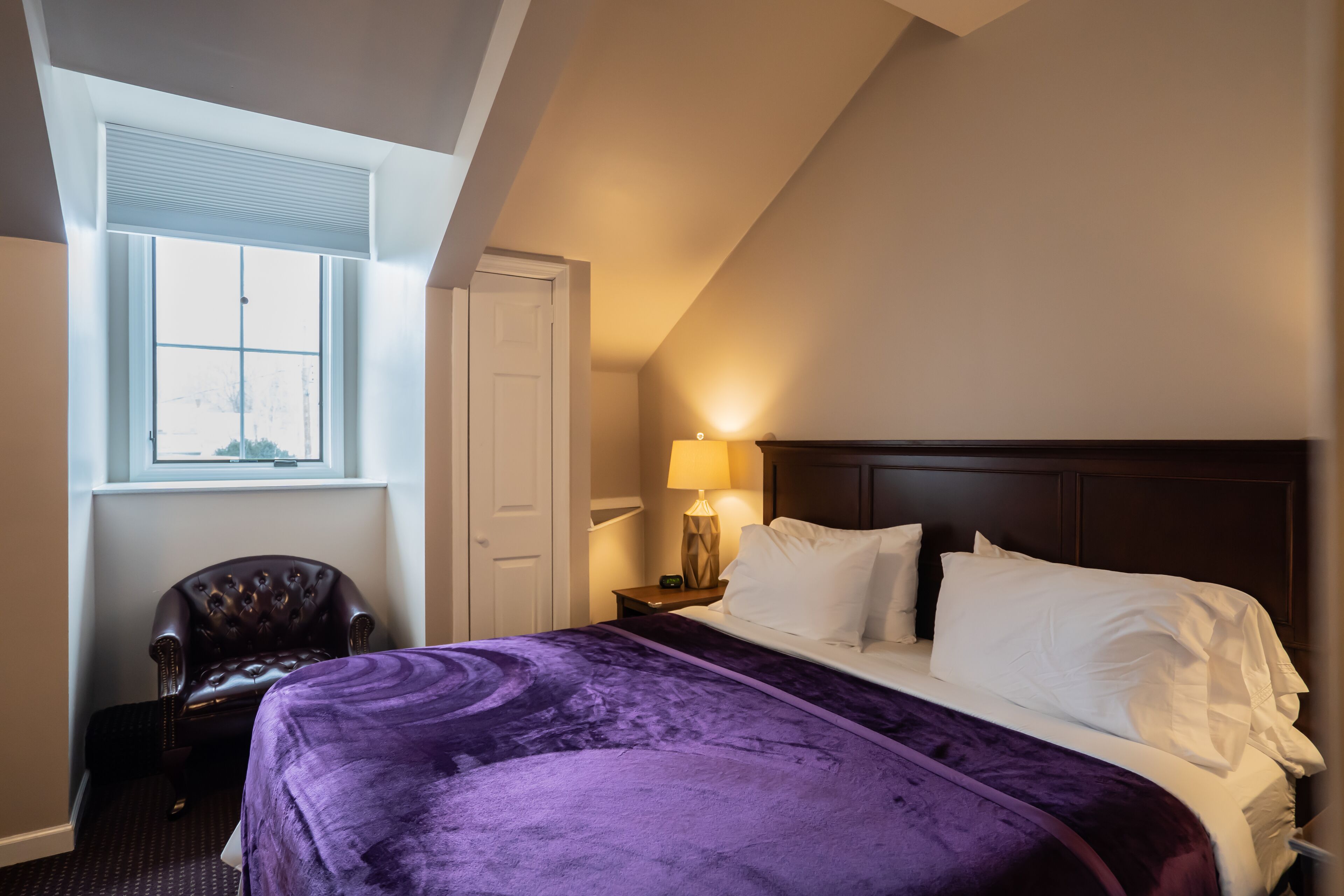 A hotel room with a large bed, a chair, a nightstand with a lamp, and a window with blinds.