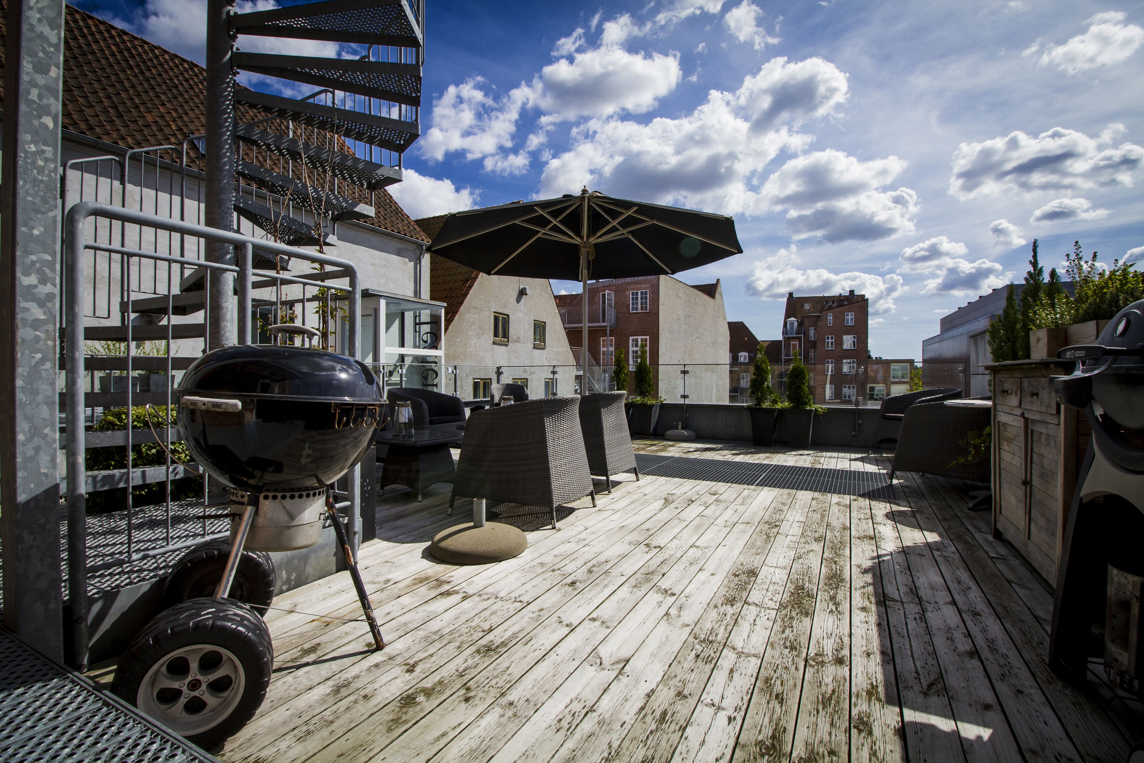 Una terraza de madera con mobiliario exterior, una parrilla y un gran paraguas.