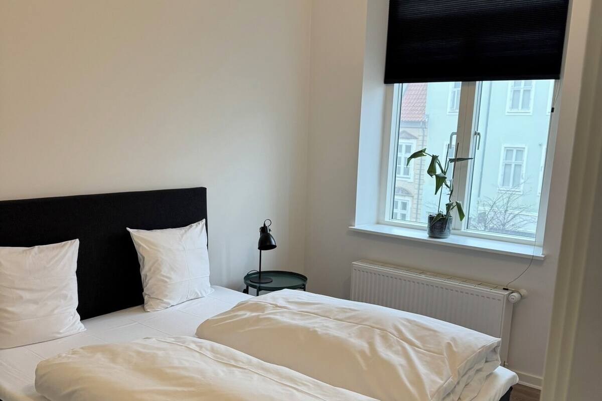 A neatly made bed with white linens in a room with a black headboard, a small bedside table with a lamp, and a window with a view of buildings outside.