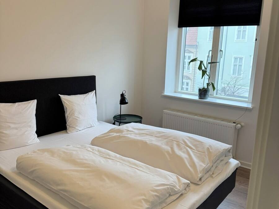 A neatly made bed with white linens in a room with a black headboard, a small bedside table with a lamp, and a window with a view of buildings outside.