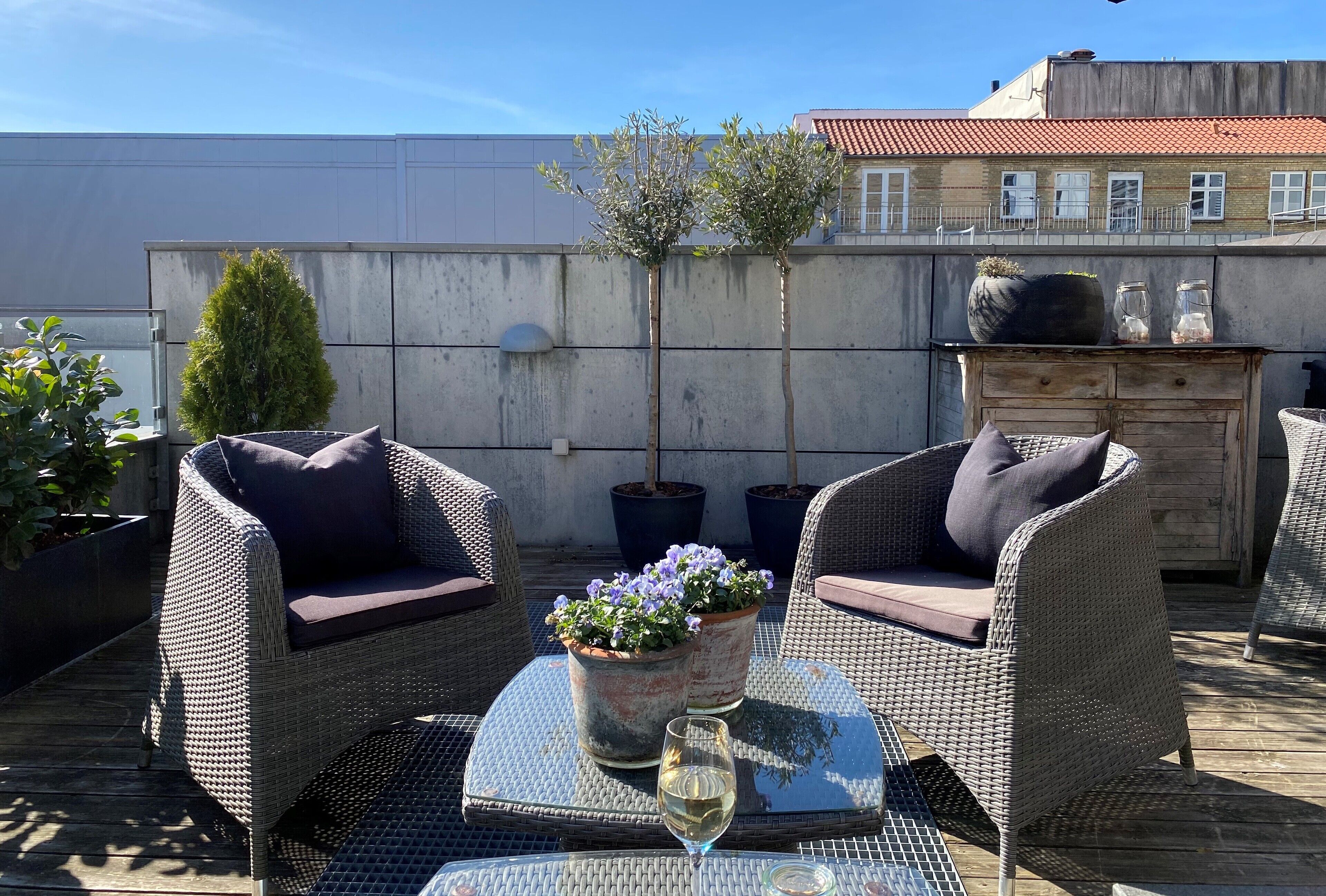 A rooftop patio with wicker furniture, a glass table, and potted plants.
