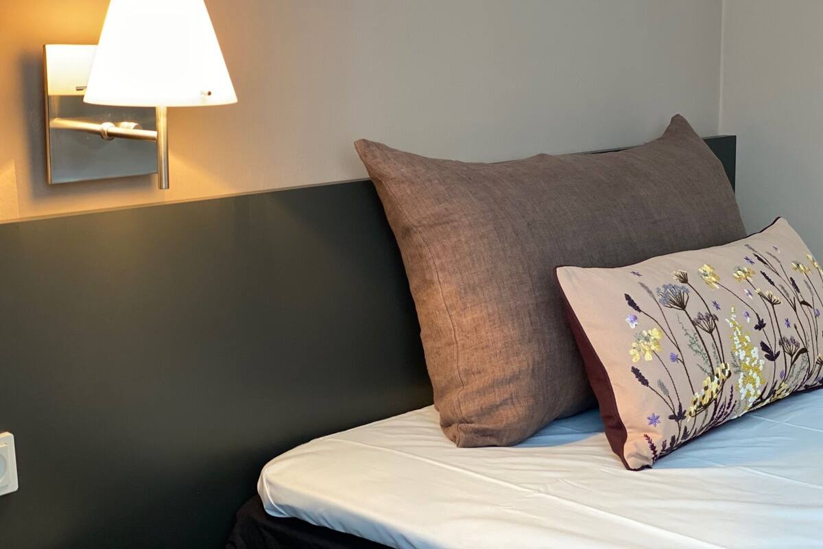 A neatly made bed with a white mattress and two pillows, one plain and one with a floral design. A wall-mounted lamp provides lighting.