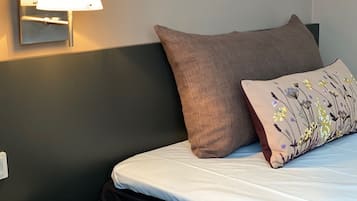 Una cama bien hecha con un colchón blanco y dos almohadas, una lisa y otra con diseño floral. Una lámpara de pared proporciona iluminación.