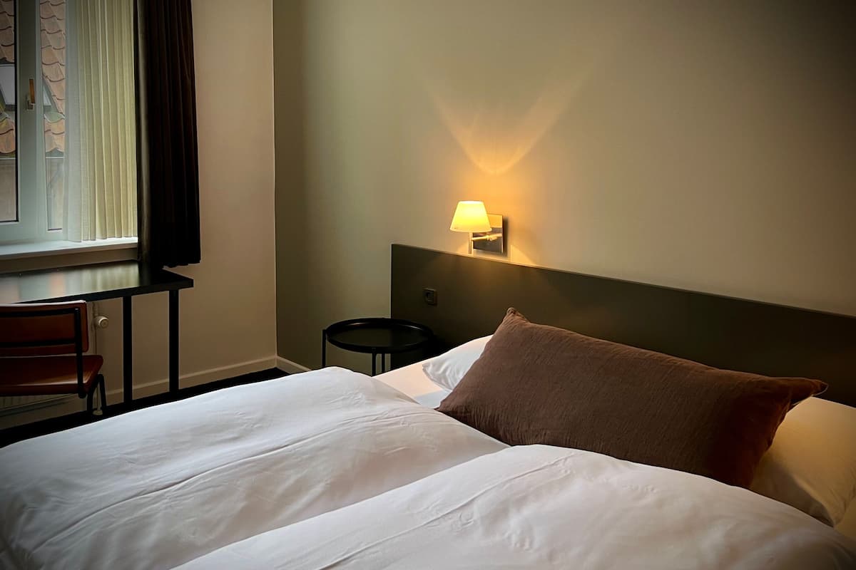 A hotel room with a large bed, a nightstand, a lamp, a chair, and a window with blinds.