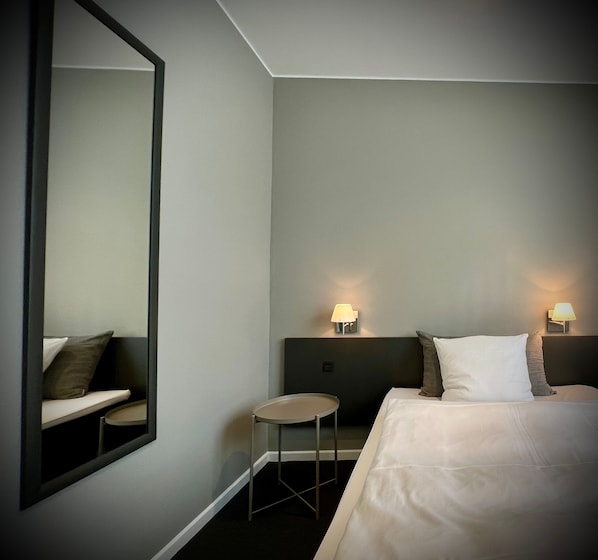 Una habitación de hotel con una cama, una mesa redonda, una silla, un espejo grande y dos lámparas fijadas a la pared.