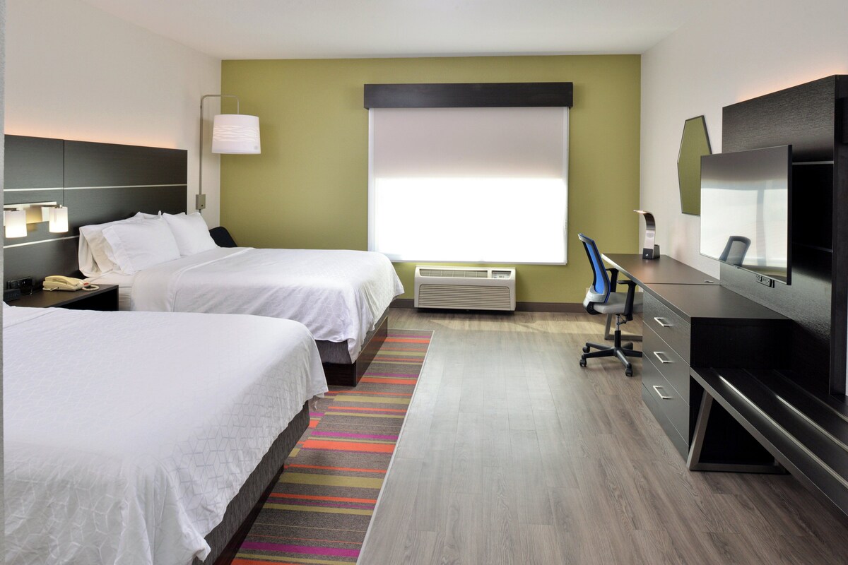 Ein Hotelzimmer mit zwei Betten, einem Schreibtisch mit Stuhl, einem Fernseher und einem Fenster mit Jalousien.