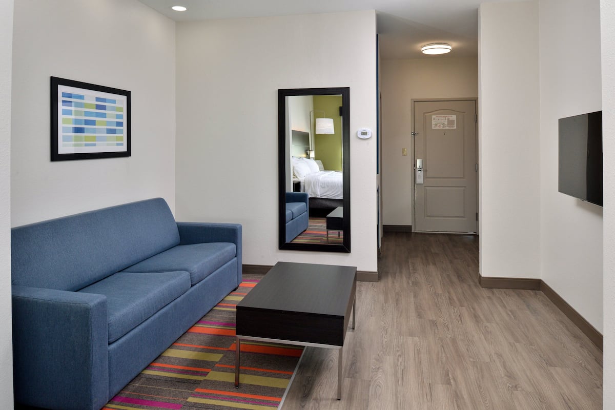 Une chambre d’hôtel moderne avec un canapé bleu, une petite table noire, un tapis rayé aux couleurs vives, un miroir et un téléviseur fixé au mur.