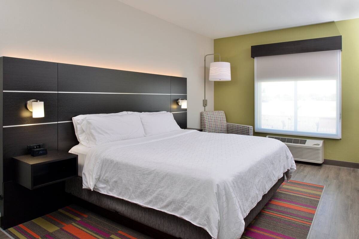 A modern hotel room with a large bed, a nightstand with a clock, a wall-mounted lamp, and a window with blinds.
