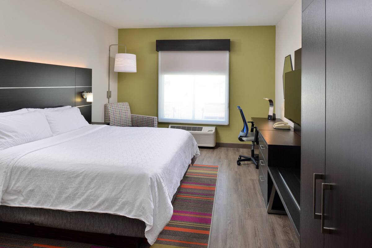 A hotel room with a large bed, a desk, a chair, a window with blinds, a lamp, and a TV.
