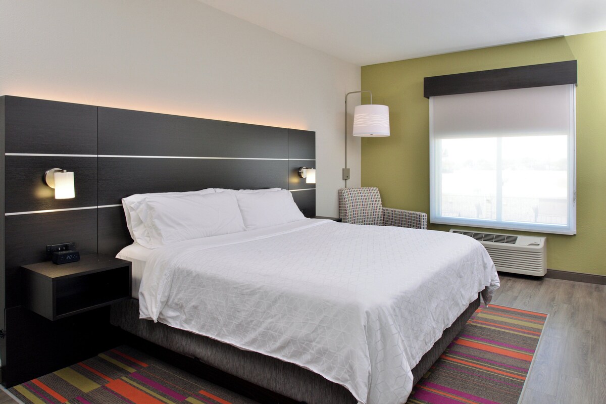 Ein modernes Hotelzimmer mit einem großen Bett, einem Nachttisch mit einer Uhr, einer Wandlampe und einem Fenster mit Jalousien.