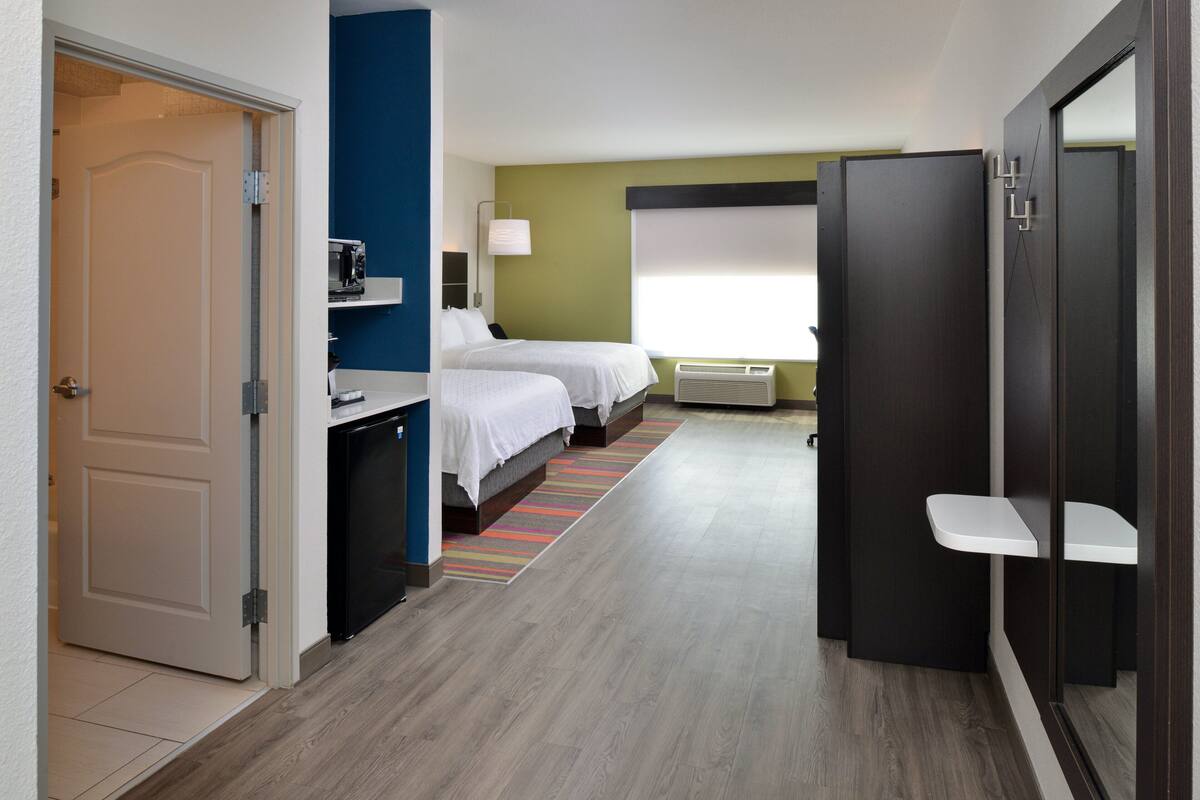 A hotel room with two beds, a desk, a chair, a small table, a window with blinds, and a door.