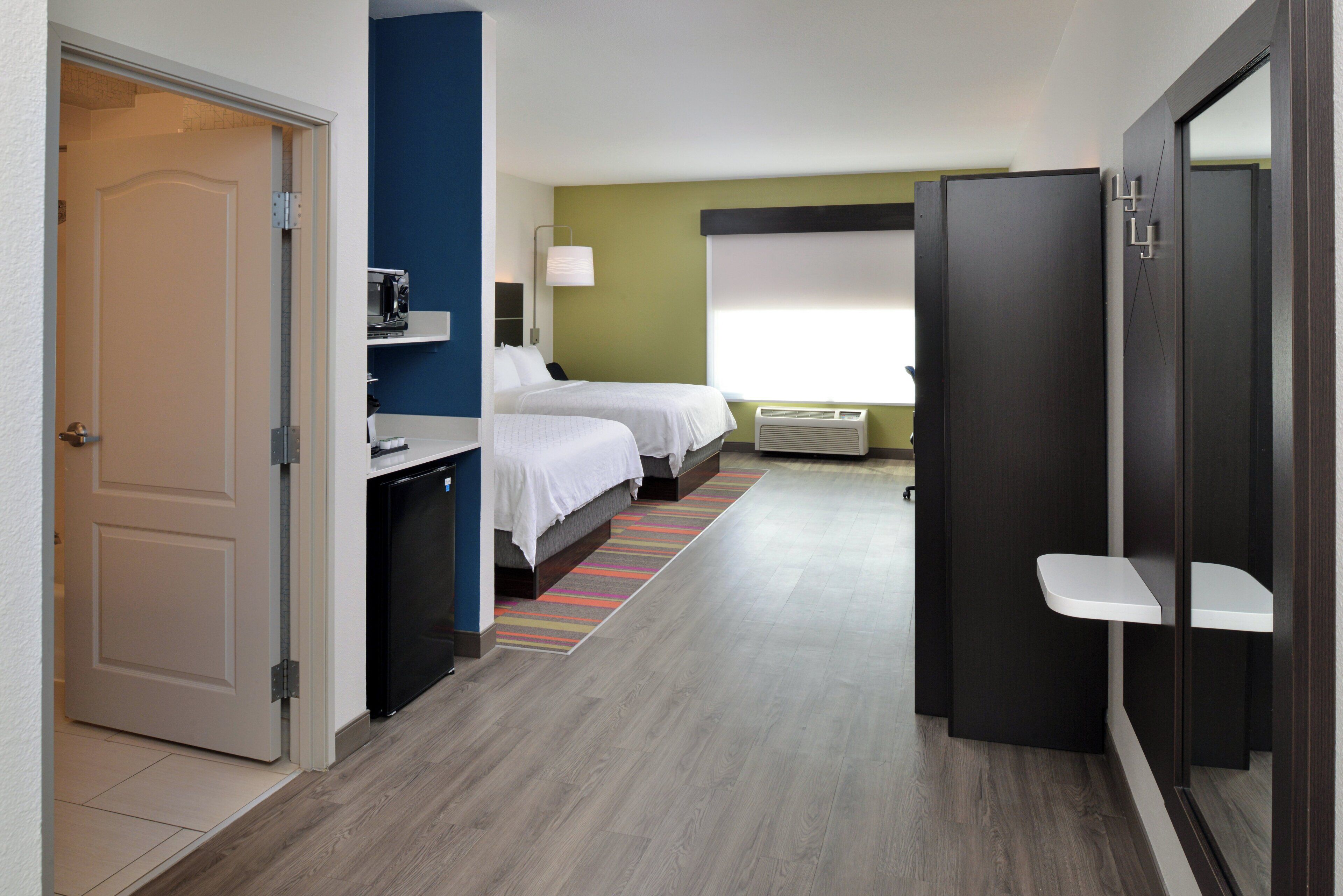 A hotel room with a bed, a desk with a microwave, a window with blinds, and a door.