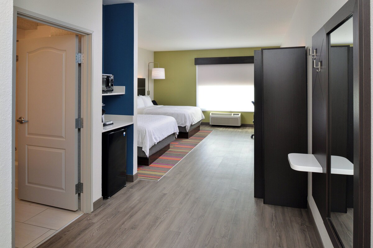 Ein Hotelzimmer mit zwei Betten, einem Schreibtisch, einem Stuhl, einem kleinen Tisch, einem Fenster mit Jalousien und einer Tür.
