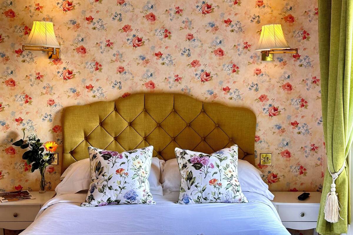 Une chambre à coucher avec un papier peint à motifs floraux, une tête de lit capitonnée, deux lampes de chevet, un rideau vert et un vase rempli de fleurs posé sur la table de chevet.