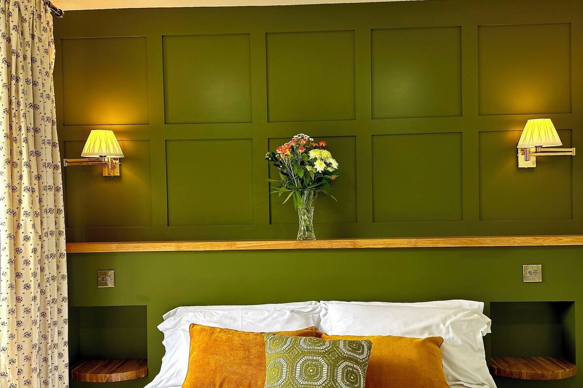 Un lit bien fait, agrémenté d’oreillers blancs et moutarde, d’un coussin à motifs verts et d’un vase de fleurs posé sur une étagère en bois.