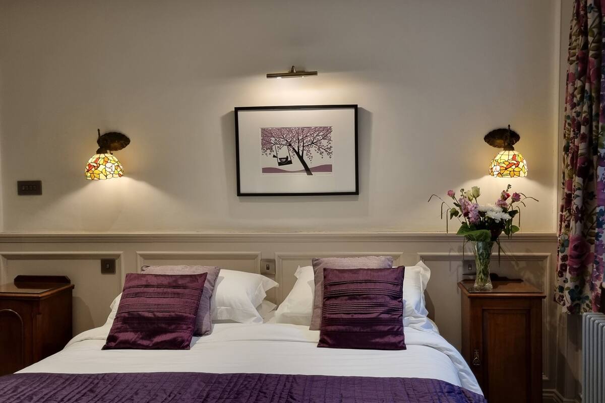 Une chambre d’hôtel avec un lit, deux tables de chevet, un tableau encadré, un vase de fleurs et une fenêtre avec des rideaux.