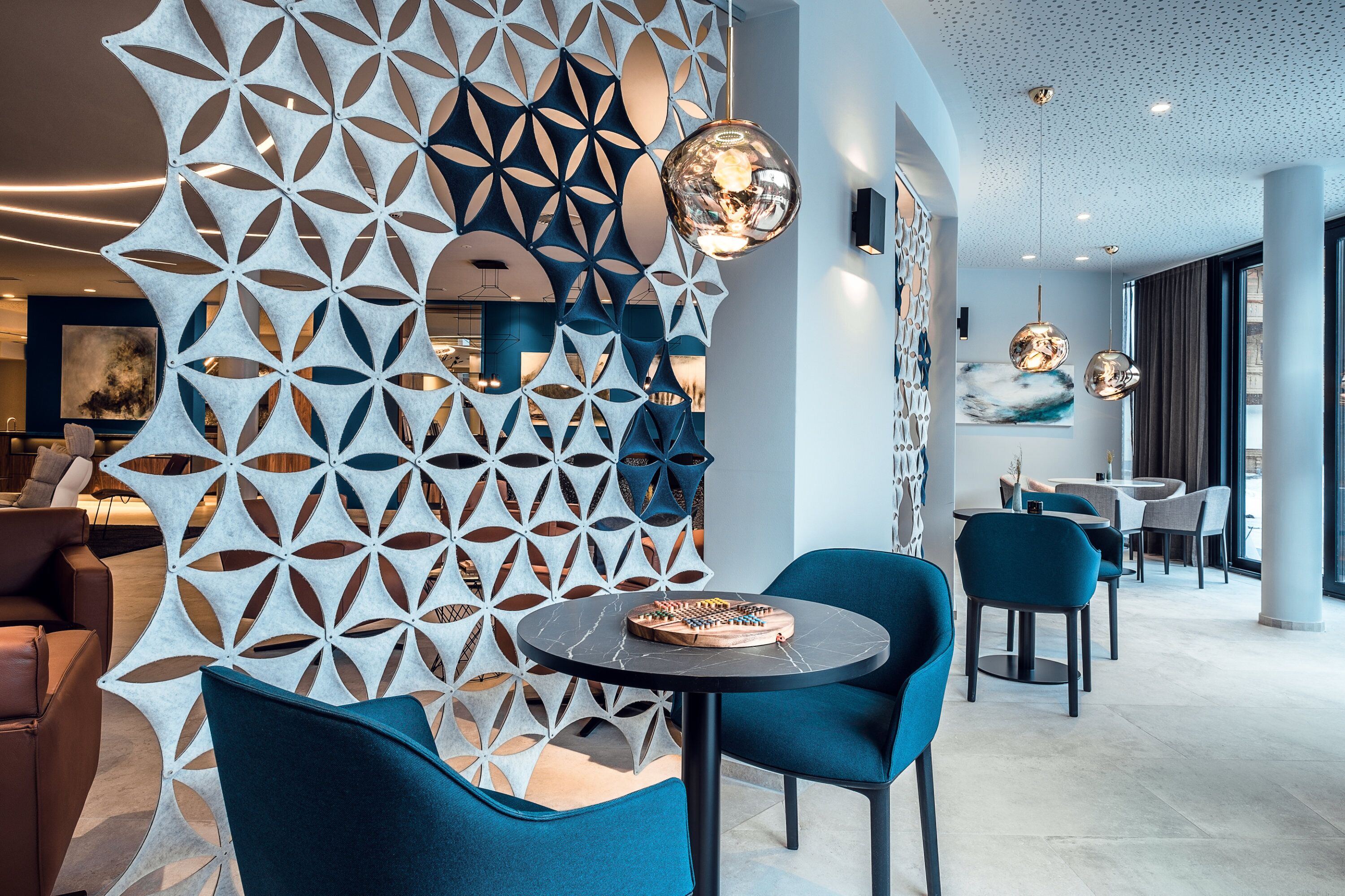 Un intérieur moderne avec un motif géométrique sur le mur, une table ronde sur laquelle est posé un jeu de société, et des chaises bleues.