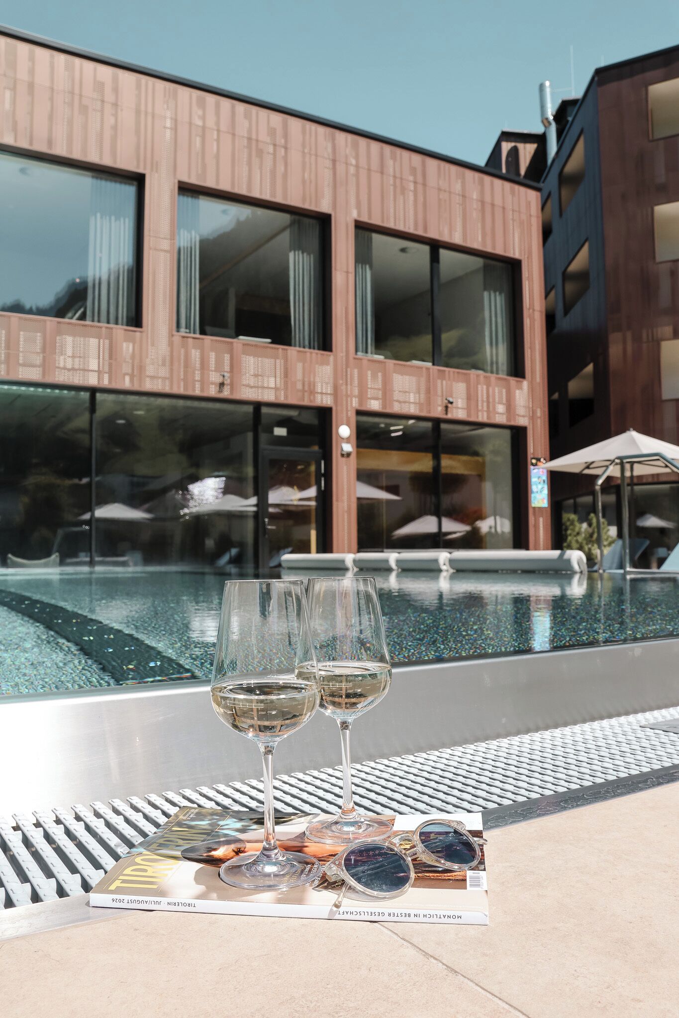 A poolside setting with two glasses of white wine on a tray, a magazine, and sunglasses.