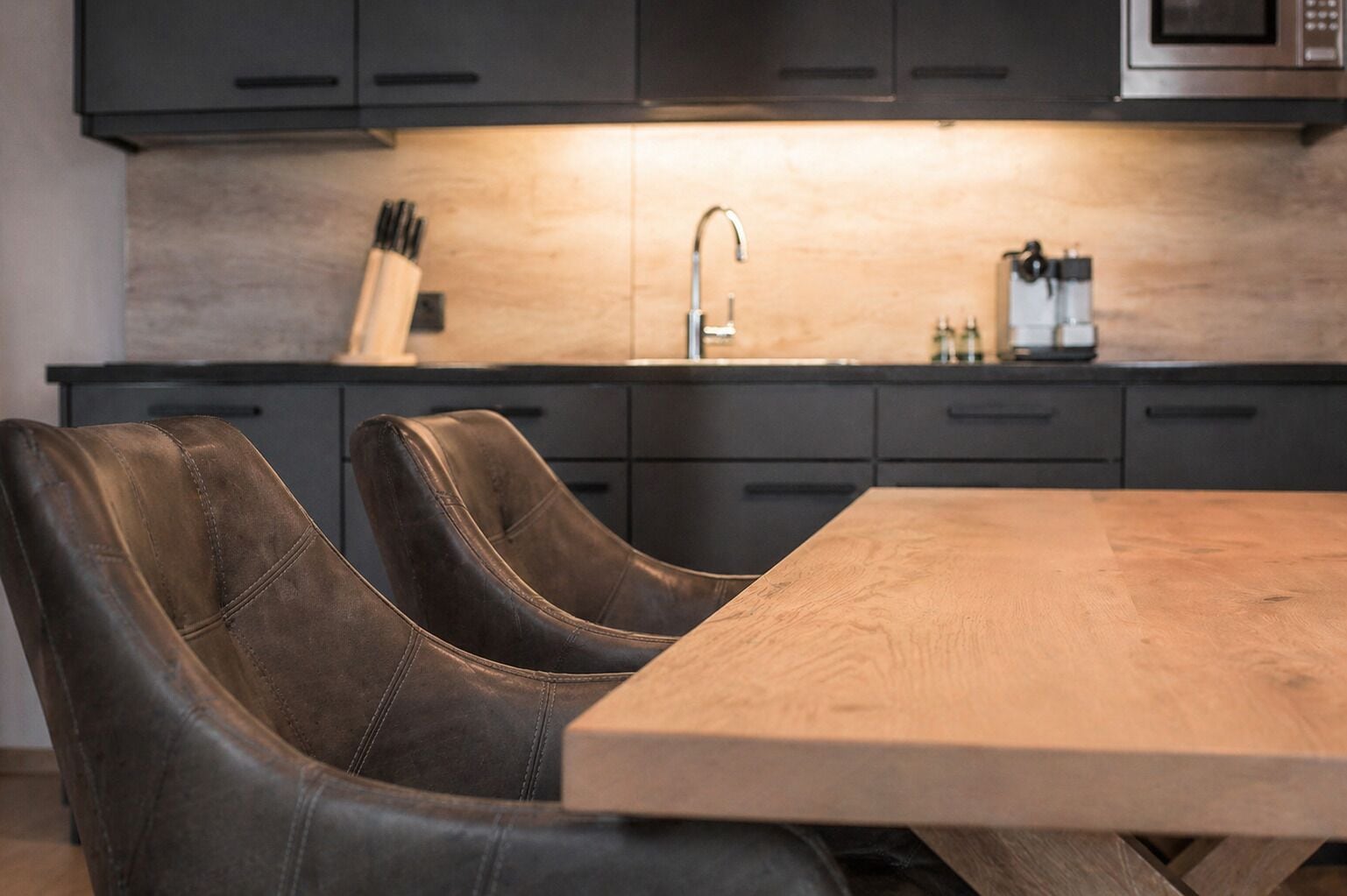 A modern kitchen with dark cabinets, a wooden dining table, and two leather chairs.