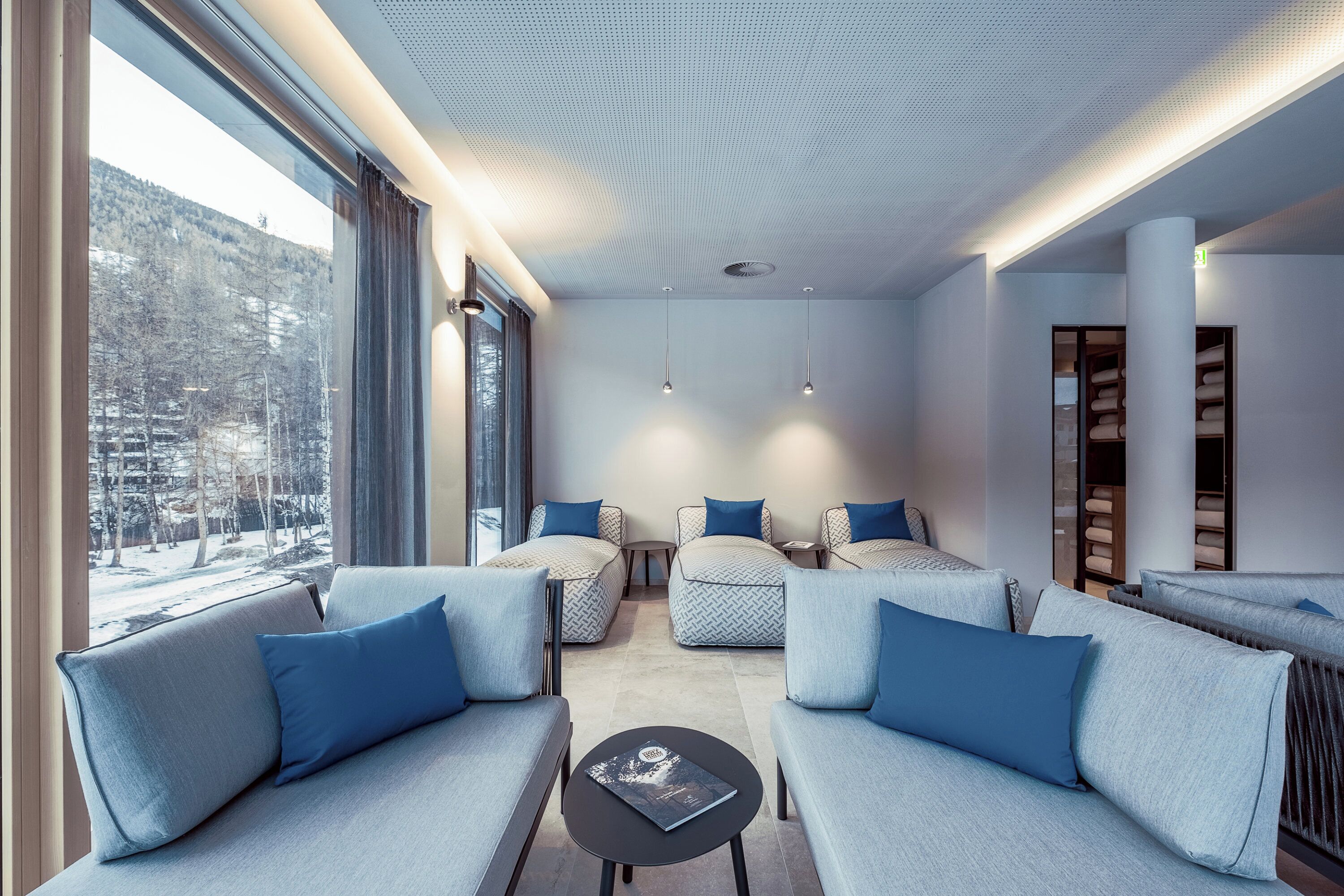 A modern living room with sofas and armchairs, a coffee table, pendant lights, and a view of a snowy landscape through large windows.