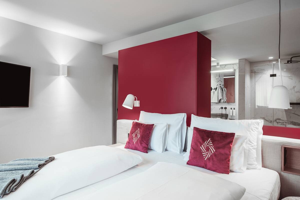 A modern hotel room with a large bed, red accent wall, and a bathroom with a marble shower.