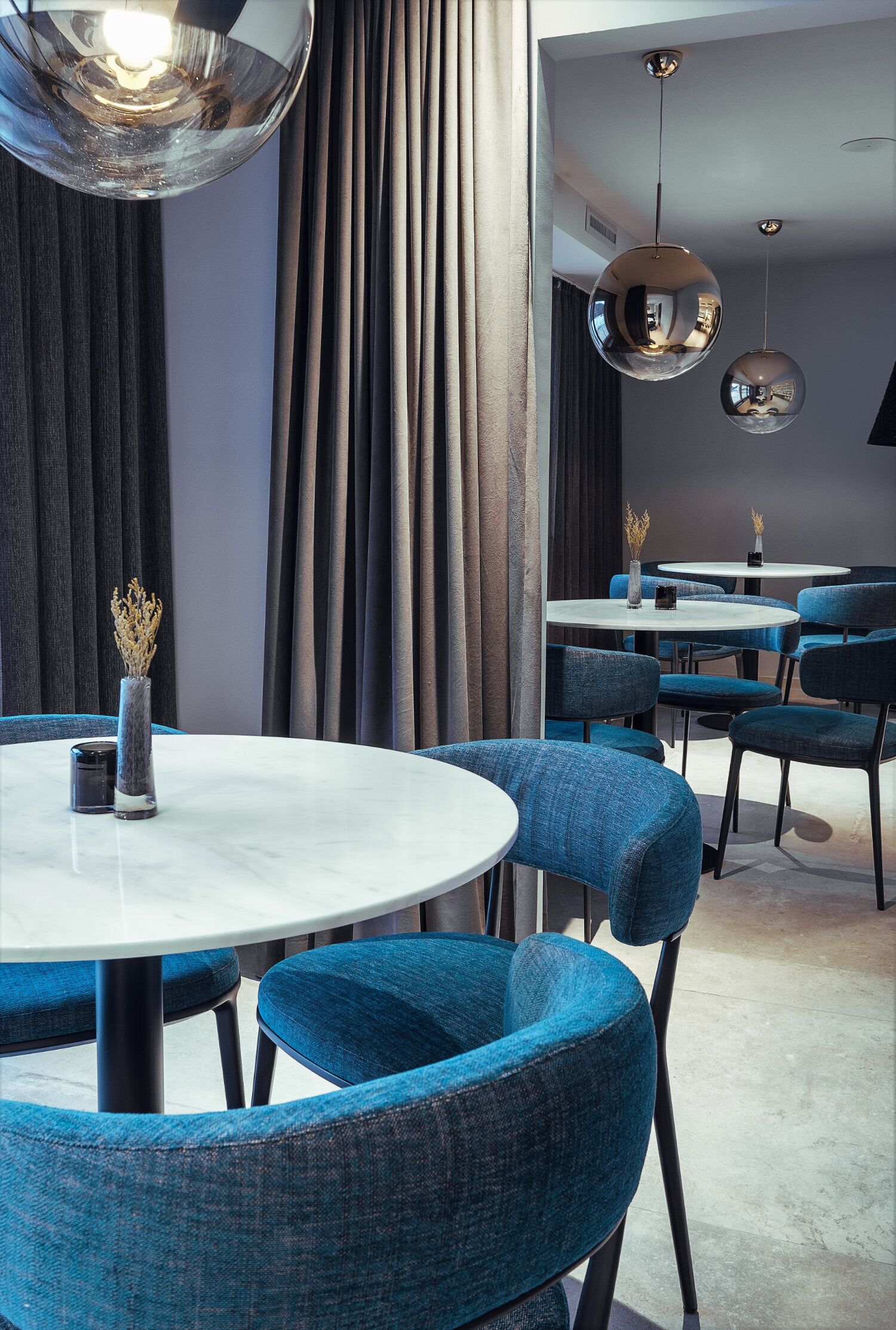 Un comedor moderno con mesas redondas y sillas tapizadas en azul, con lámparas colgantes reflectantes y un jarrón con plantas secas.