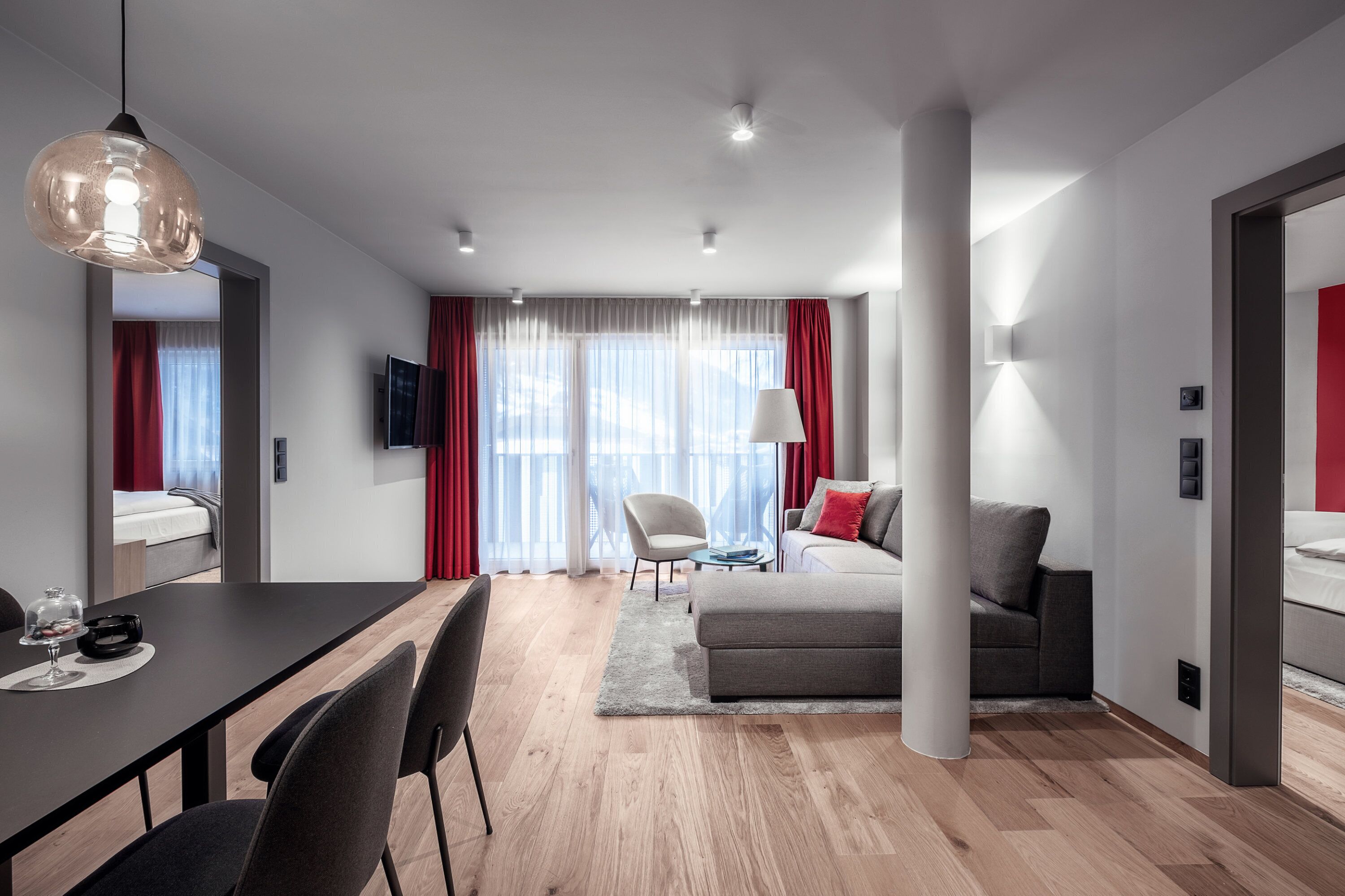 A modern hotel room with a dining area, a sofa, a TV, and a large window with curtains.
