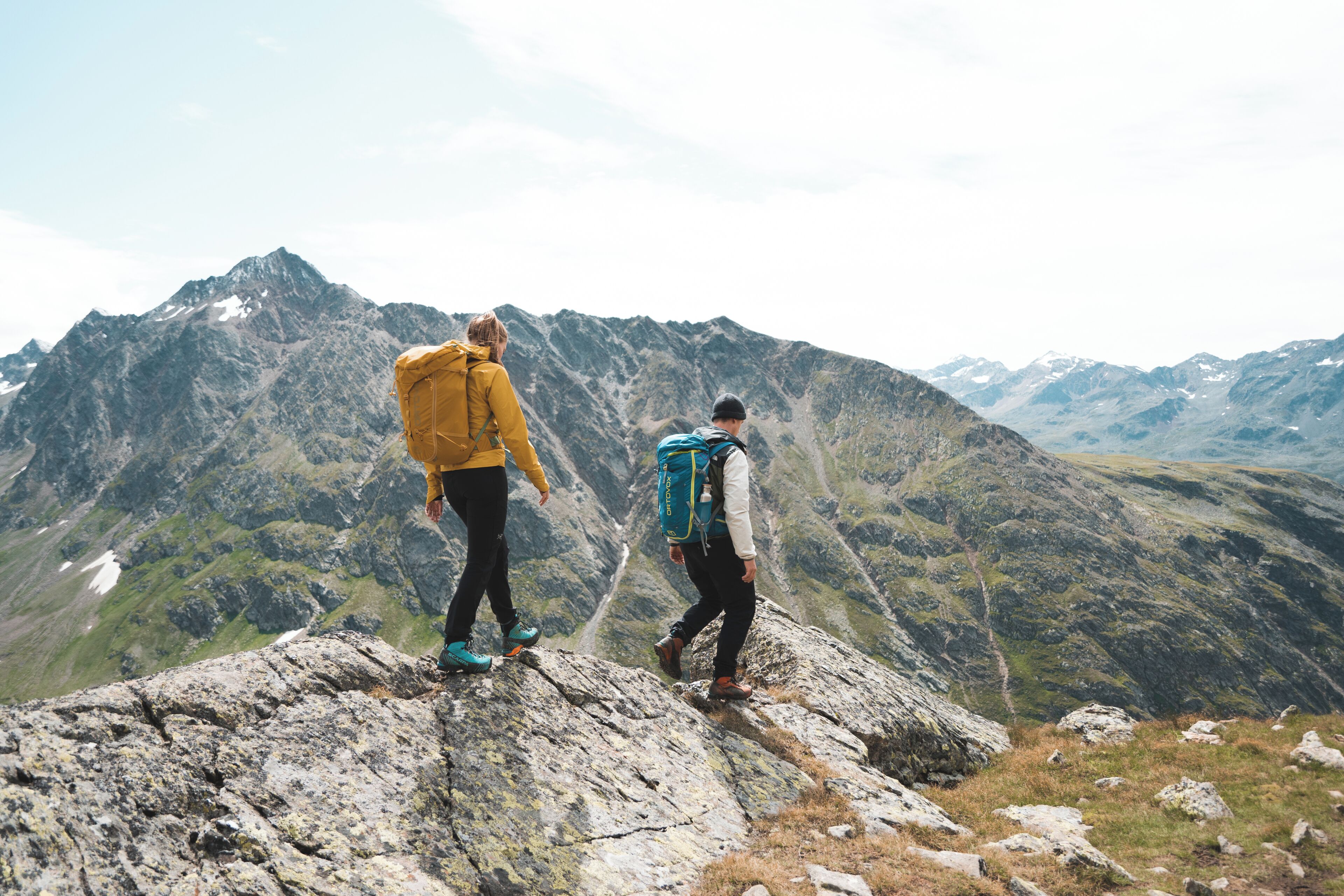 Deux randonneurs se trouvent sur une crête montagneuse, avec une chaîne de montagnes en arrière-plan.