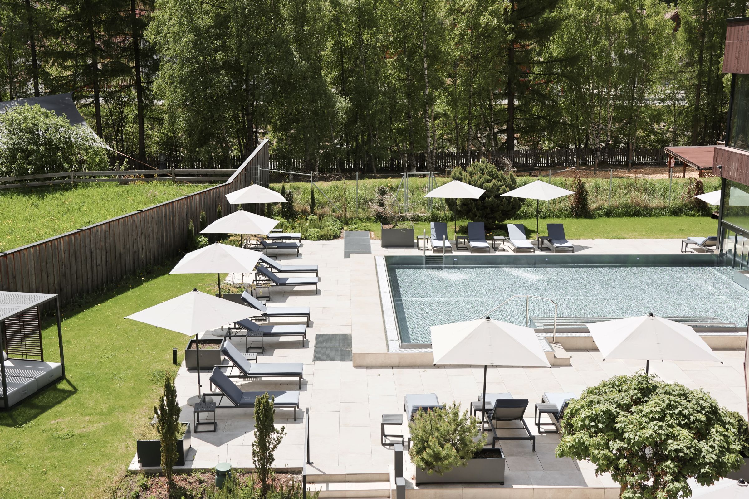 A rooftop pool area with lounge chairs and umbrellas, surrounded by a wooden fence and greenery.