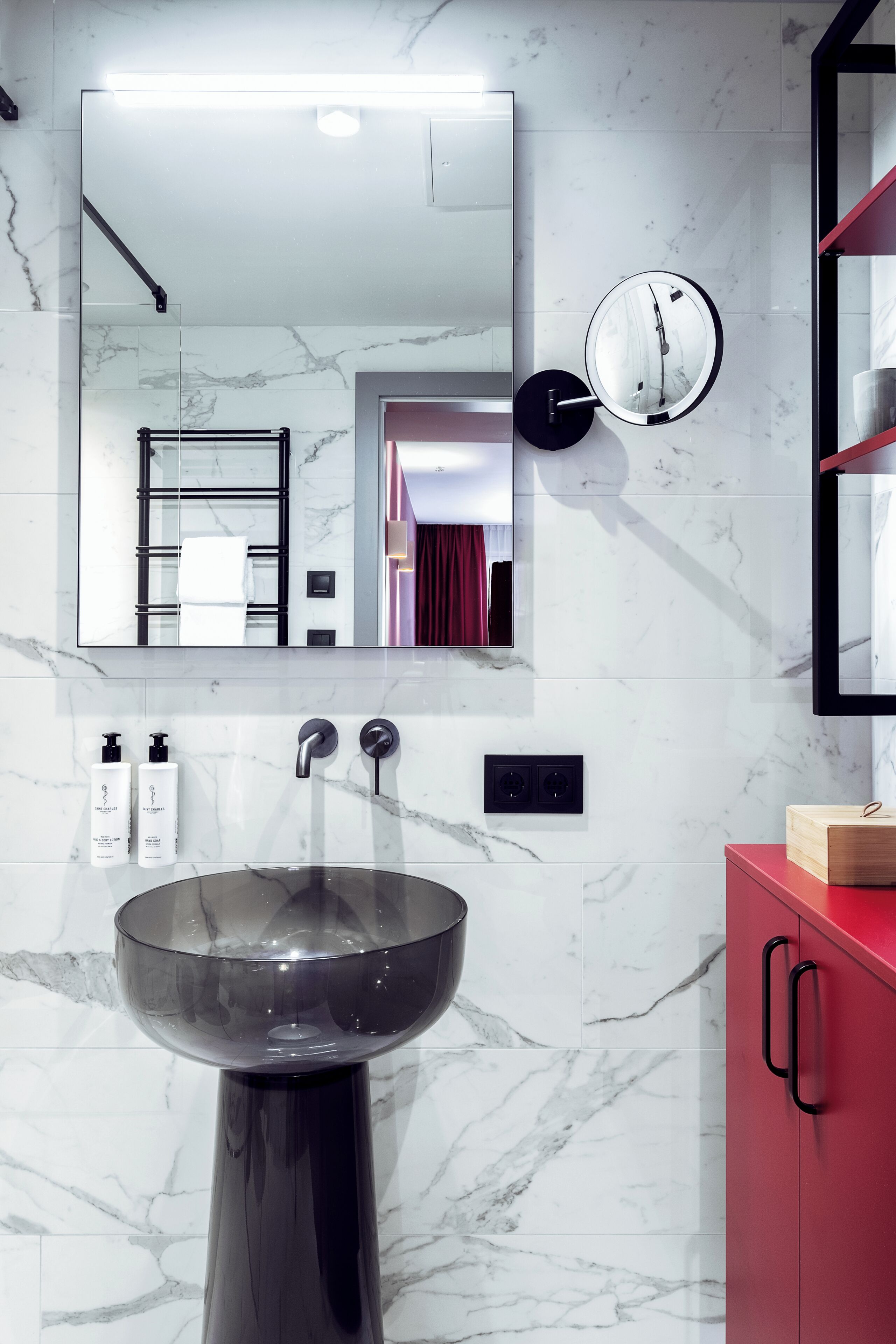 Un baño moderno con un espejo grande, un reloj redondo en la pared, un lavabo de vidrio y un mueble rojo.