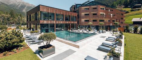 A modern hotel with a swimming pool, lounge chairs, and a mountainous backdrop.