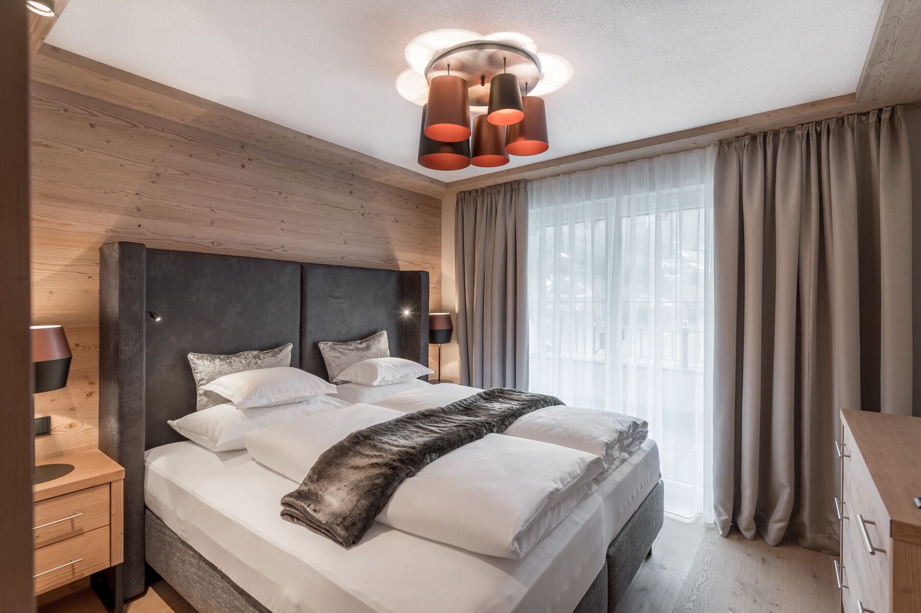 A modern hotel room with a large bed, a wooden headboard, a ceiling light fixture, and a window with curtains.