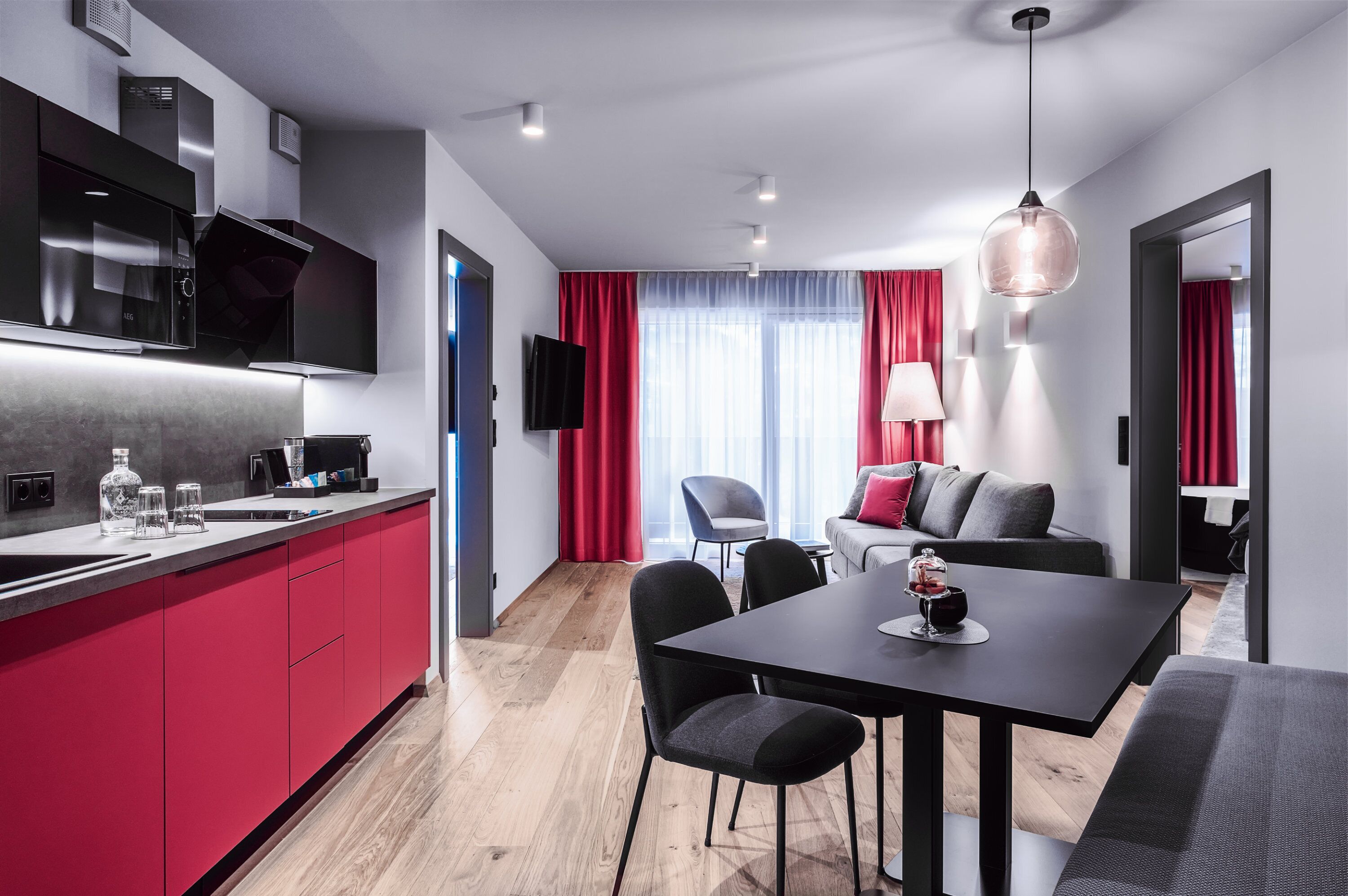 A modern kitchen with red cabinets, a dining area with a black table, and a living room with a gray sofa.