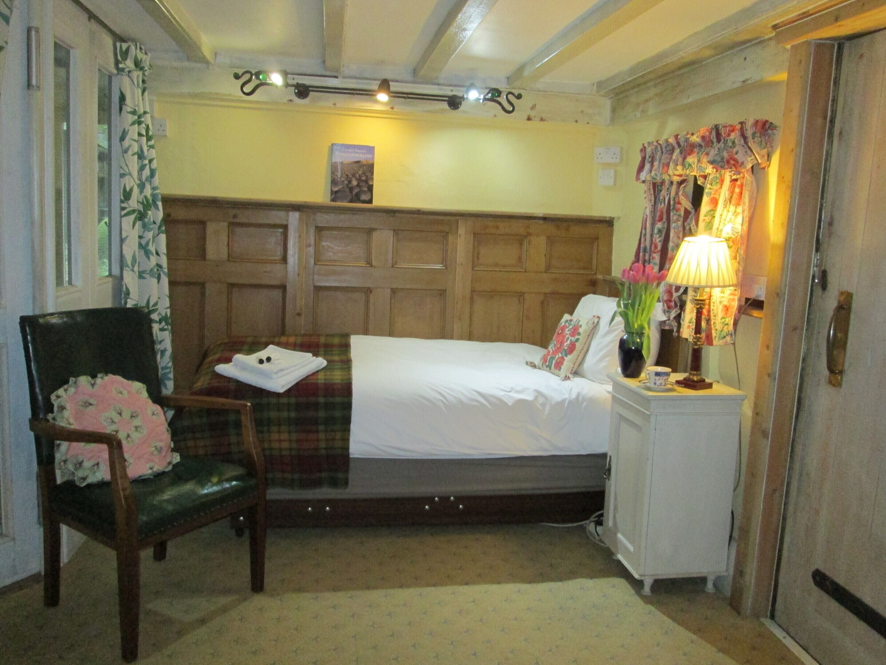 Une chambre à coucher avec une tête de lit en bois, un lit simple, une chaise avec un coussin à motifs floraux, une table de chevet avec une lampe et une petite armoire.