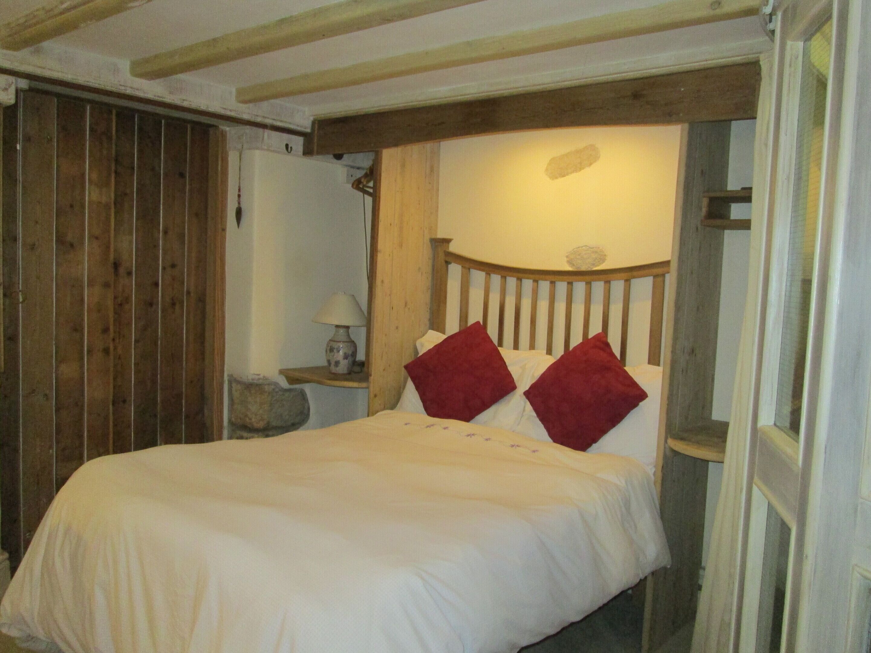 A bed with a wooden headboard and two red pillows, a bedside table with a lamp, and a stone wall.