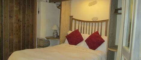 A bed with a wooden headboard and two red pillows, a bedside table with a lamp, and a stone wall.