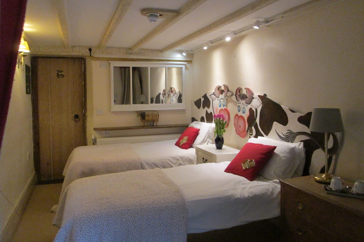 Quarto de hotel com duas camas, teto de madeira, espelho e um quadro de vaca na parede.