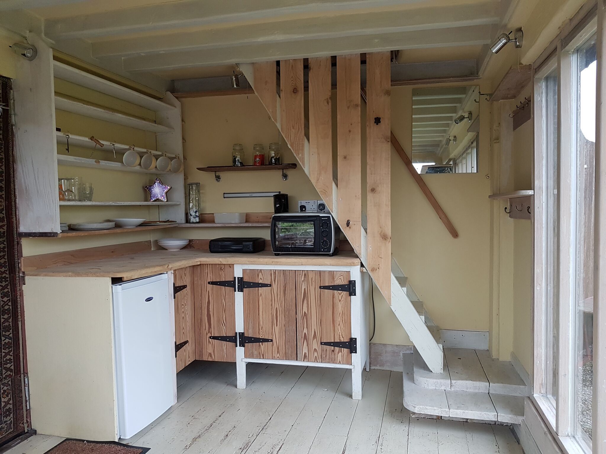 Une cuisine compacte avec des meubles en bois, un four à micro-ondes encastré et un petit réfrigérateur. Un escalier en bois mène à une porte située sur le côté droit.