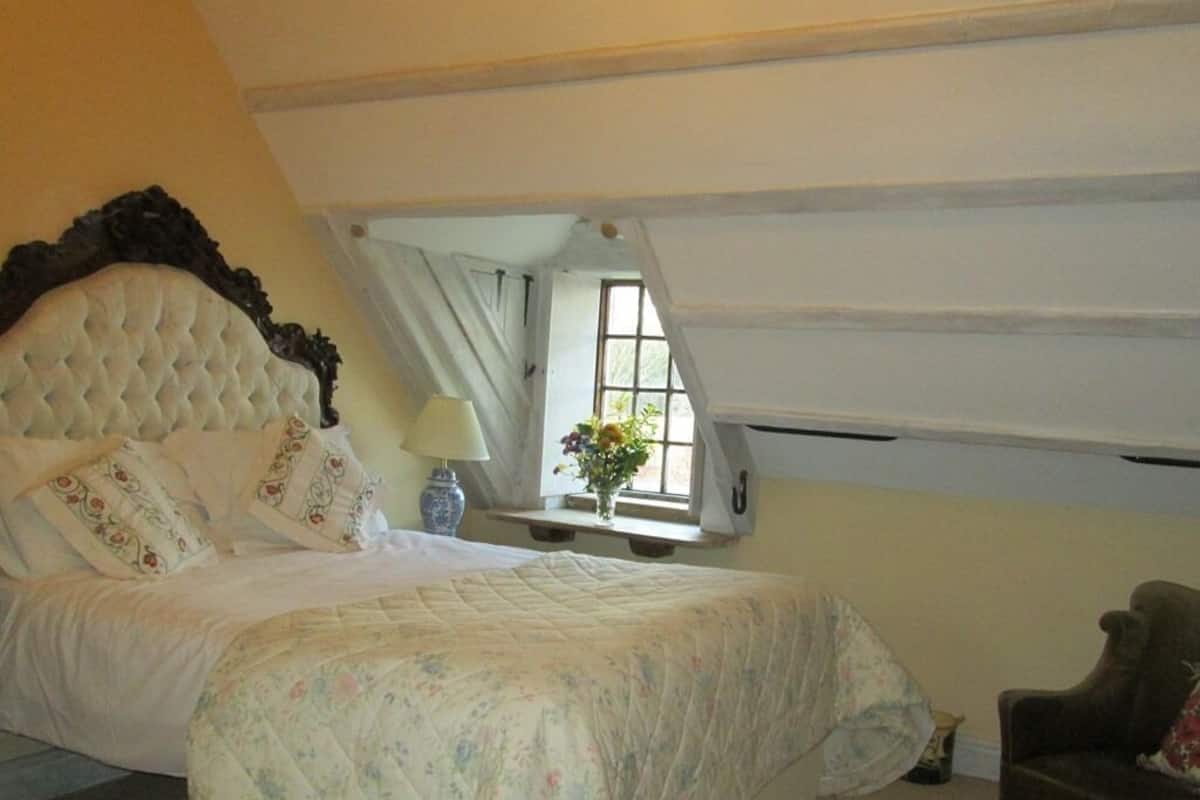 Quarto com cama grande, janela com cortinas e pequena mesa com um vaso de flores.
