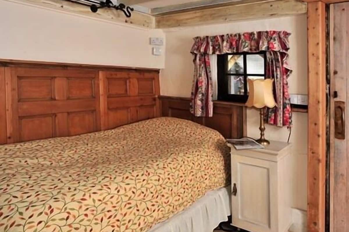 Quarto com cabeceira de madeira, colcha estampada, mesa de cabeceira com abajur e janela com cortinas florais.