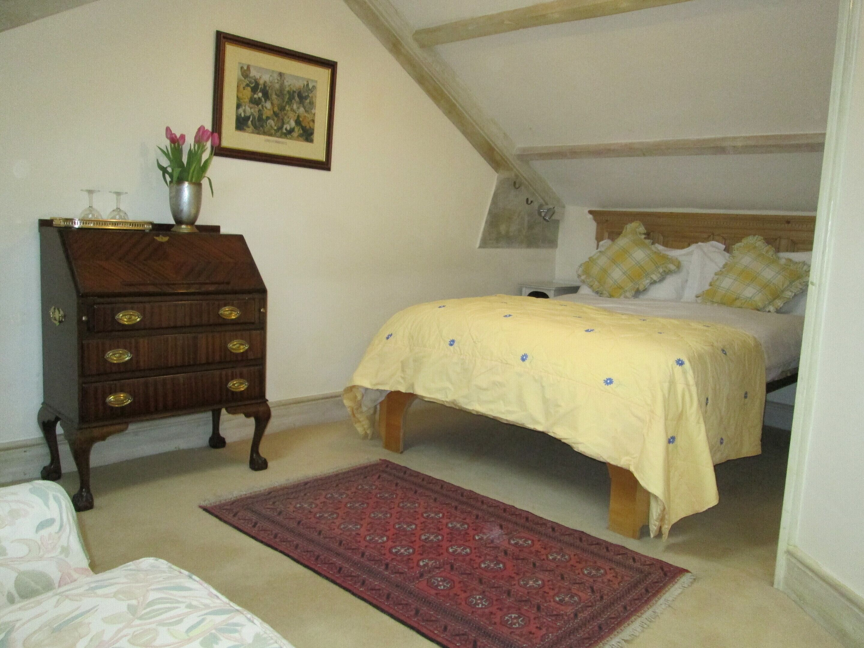 A bedroom with a bed, a wooden dresser, a red rug, a painting, and a vase with flowers.