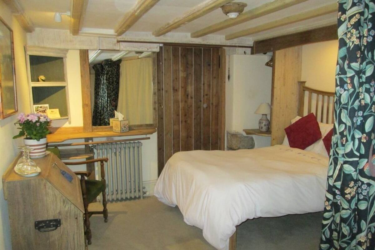 Quarto com cama de madeira, uma escrivaninha com cadeira, radiador e janela com cortinas.