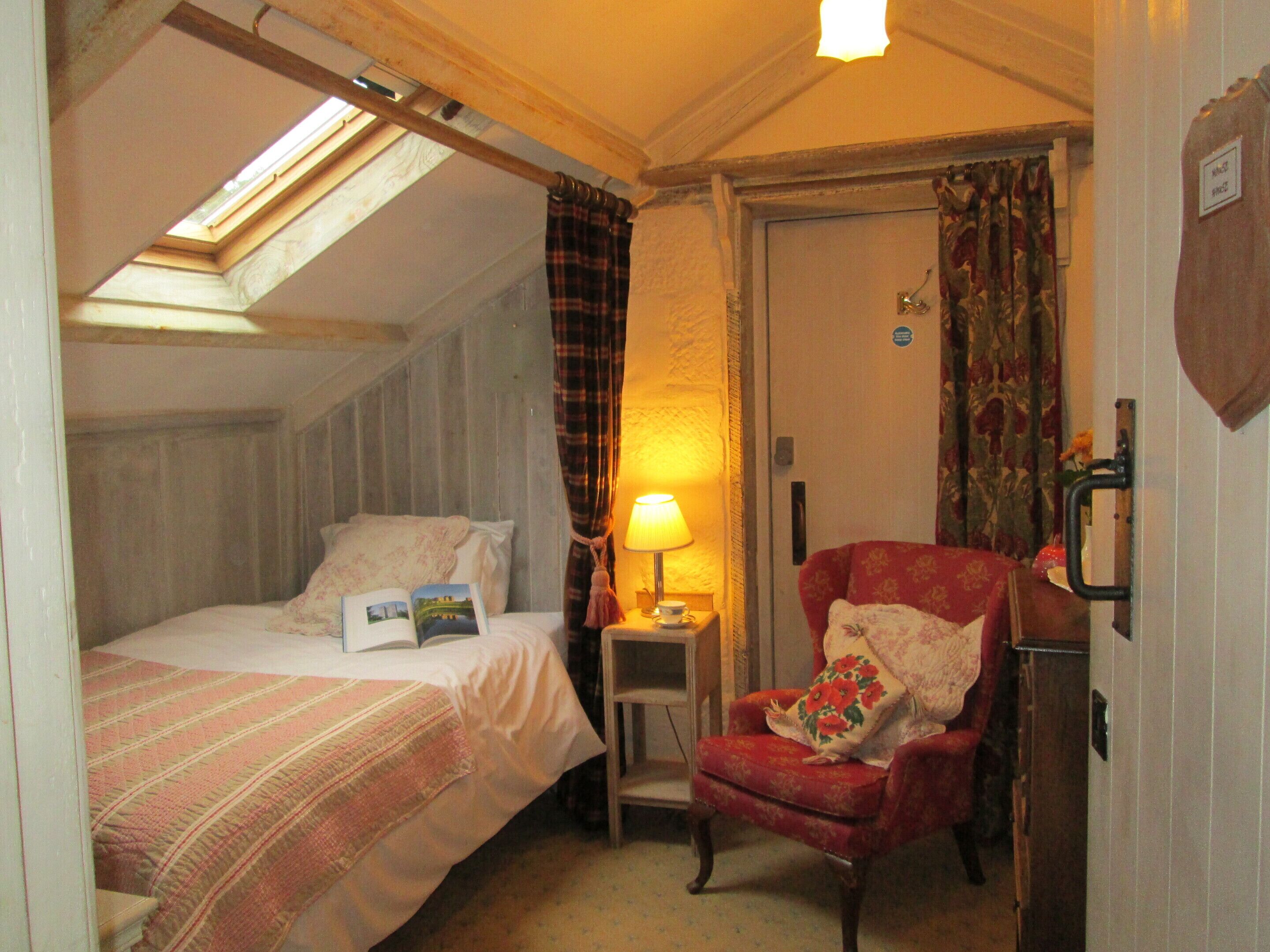 A bedroom with a bed, a reading chair, a nightstand with a lamp, a door, and a window with curtains.